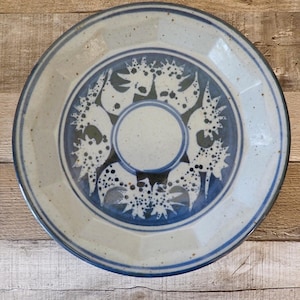May include: A ceramic plate with a light beige rim and a blue and white patterned center. The center design features a circular pattern with stylized floral or abstract shapes. The plate has a diameter of approximately 10 inches.