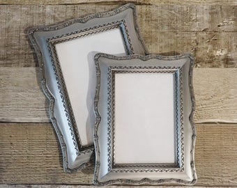 May include: Two rectangular picture frames with a metallic silver finish. Each frame features an ornate, raised border with a repeating floral pattern and a delicate scalloped inner edge. The frames are empty, displaying white rectangular centers.
