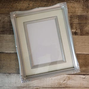 May include: Silver-colored picture frame with ornate detailing on the corners. The frame has a light-colored mat and a smaller inner frame. The frame is empty, ready to display a photograph or artwork.