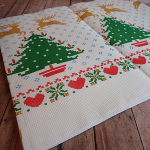 May include: Two folded white Christmas-themed towels with a pixelated design. The towels feature a green Christmas tree, red hearts, and gold reindeer. The design is in a cross-stitch style, with a textured waffle weave.