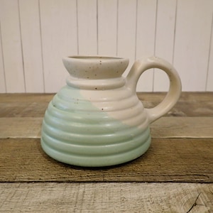 May include: A ceramic mug with a wide, rounded base and a narrow neck, featuring a ribbed, horizontal texture. The mug is finished in a speckled cream color on the upper portion and a soft, muted sage green on the lower half, with a curved handle.