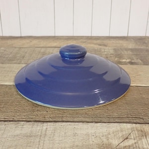 May include: A circular, deep blue ceramic lid with a smooth, glossy finish. The lid features a stepped, concentric design that slopes gently toward a small, rounded knob handle at the center. The underside edge shows a light, unglazed rim.