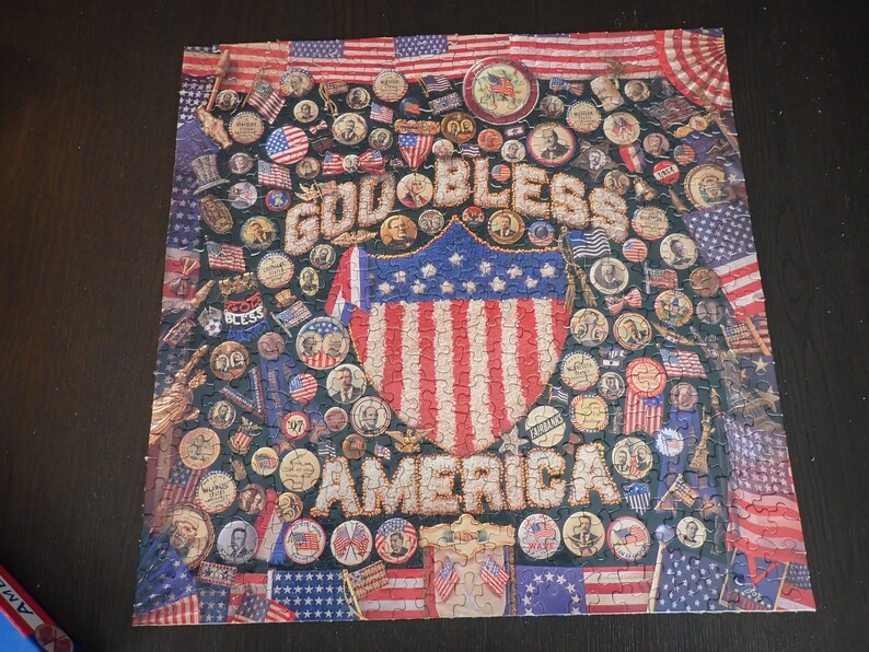 Może przedstawiać: Ukończone puzzle o tematyce patriotycznej. Puzzle przedstawiają słowa "GOD BLESS AMERICA" w ozdobnej czcionce, otoczone obrazami amerykańskich flag, pieczęci patriotycznych i portret&oacute;w postaci historycznych. Schemat kolor&oacute;w to czerwony, biały i niebieski.