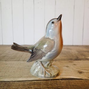 May include: A ceramic bird figurine with a light blue head, orange chest, and gray wings and tail. The bird is perched on a gray base and is looking upwards. The figurine is on a wooden surface.