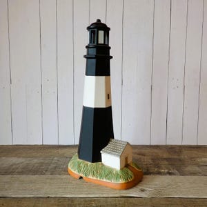 May include: A lighthouse figurine featuring a tall, tapered tower with alternating black and white horizontal bands, a black lantern room at the top, and a small white building with a textured roof attached to the base, which is painted green and brown.