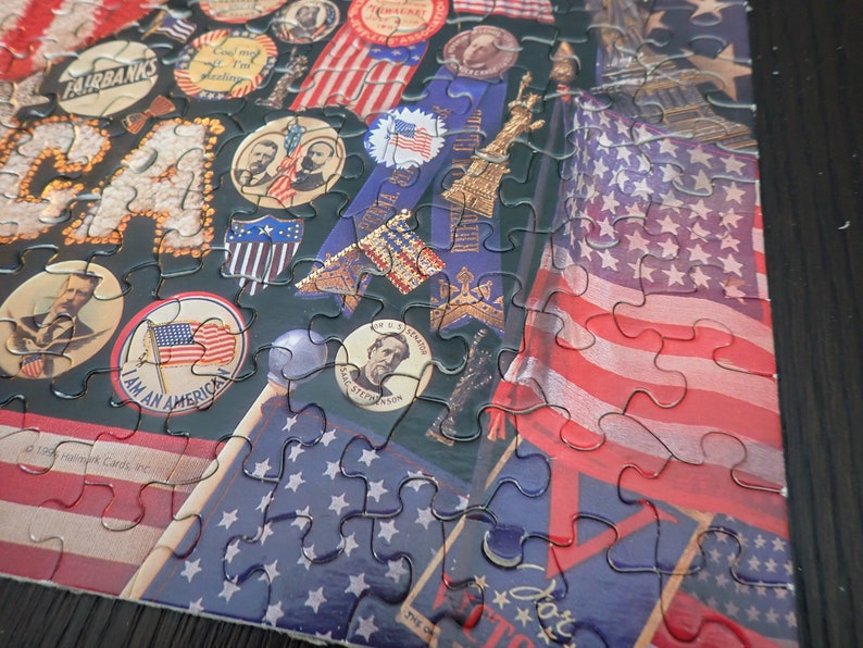 Może przedstawiać: Ukończone puzzle o tematyce patriotycznej. Przedstawiają amerykańską flagę, postacie historyczne i patriotyczne wstążki. Elementy są ze sobą połączone, tworząc kompletny obraz na ciemnym tle. Idealne na prezent.
