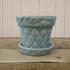 May include: A light blue ceramic plant pot with a matching saucer. The pot features a quilted diamond pattern on the body and a raised leaf motif around the rim. The saucer also displays a matching raised leaf design along its outer edge.
