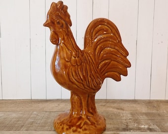 May include: A brown ceramic rooster figurine. The rooster is standing upright with its head turned to the left. The figurine has a glossy finish and is set on a round base. The rooster's comb, wattle, and tail feathers are detailed.