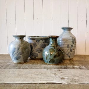 May include: A collection of four ceramic vessels including two small round vases with blue leaf patterns, one cylindrical planter with a blue leaf motif, and one taller light blue vase with a dark blue painted branch design and a narrow neck.