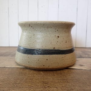 May include: A speckled, beige ceramic pot with a dark blue horizontal stripe. The pot has a slightly flared rim and a rounded base. The handmade pottery is a neutral color and would be a decorative addition to any home.