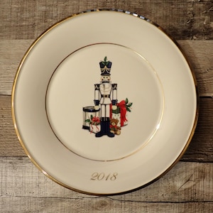 May include: A white decorative plate with a gold rim and a gold inner ring. The plate features a nutcracker soldier, a drum, wrapped gifts, and a teddy bear. The year "2018" is printed in gold on the plate.