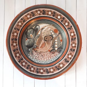 May include: A round, hand-painted ceramic plate with a detailed owl design in the center. The plate features a red rim, a floral border, and a gray and brown color scheme. The owl is depicted with intricate feather details.