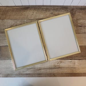 May include: A double-hinged picture frame featuring two rectangular sections with thin, metallic gold-toned borders. Each section contains a plain white insert, and the two frames are connected by a central metal hinge, allowing for an open display.