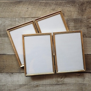 May include: A set of two hinged double picture frames with a metallic gold finish and a beaded edge detail. Each frame features a rectangular orientation with a white mat board insert, designed to hold two photographs side by side when opened.