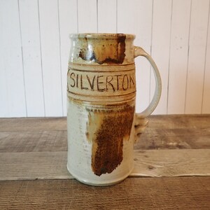 May include: A tall, handcrafted ceramic mug with a large handle. The mug is primarily beige with brown accents and the word "SILVERTON" carved into the side. The mug has a rustic, handmade appearance.