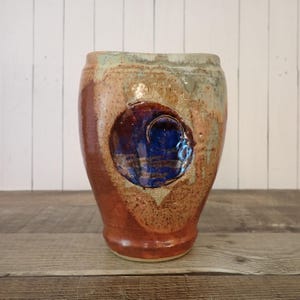 May include: A handmade ceramic vase with a unique design. The vase features a cylindrical shape with a reddish-brown base, transitioning to lighter brown and green hues towards the top. A prominent blue circular design is embedded on the front, adding a striking contrast.