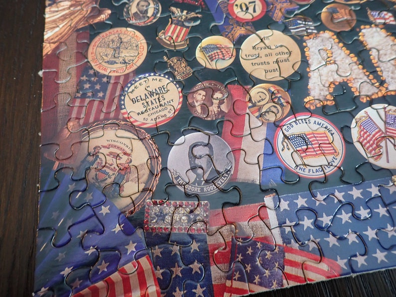 Może przedstawiać: Ukończone puzzle o tematyce patriotycznej. Elementy układanki przedstawiają amerykańskie flagi, postacie historyczne i medaliony w stylu vintage z napisami takimi jak "God Bless America" i "Theodore Roosevelt". Paleta kolor&oacute;w obejmuje czerwień, biel i błękit.
