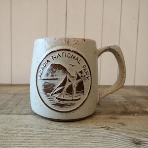 May include: A speckled off-white ceramic mug featuring a circular embossed design of a sailboat on the water with the text Acadia National Park arched above. The mug has a sturdy handle and a brown base, with a rustic, textured finish throughout.