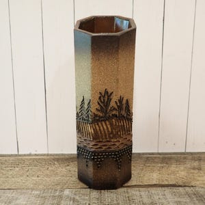 May include: A tall, hexagonal ceramic vase with a textured surface and a landscape design. The vase features a gradient of colors, transitioning from light tan to dark brown. The landscape includes trees and a textured base.