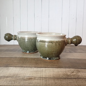 May include: Two handmade ceramic mugs with a unique handle design. The mugs have a speckled green glaze on the body, a white rim, and a dark green handle. The mugs are sitting on a wooden surface.