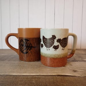 May include: Two ceramic mugs. One is brown with a leaf design, the other is white with brown chicken illustrations. The mugs have large handles and are suitable for hot beverages. The mugs are on a wooden surface.
