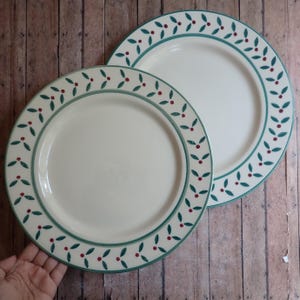 May include: Two off-white ceramic plates with a green border and a pattern of green leaves and red berries. The plates are on a wooden surface.