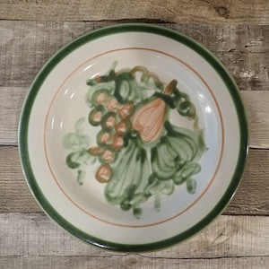 May include: A round ceramic plate with a white center, a green rim, and a thin orange line. The plate features a hand-painted design of green leaves, orange grapes, and a peach-colored fruit.