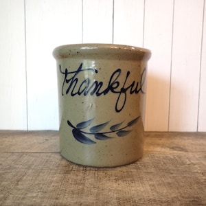 May include: A cylindrical stoneware crock with a speckled tan finish. The word Thankful is hand-painted in dark blue script across the center, positioned above a dark blue painted leafy branch motif that wraps horizontally around the vessel.