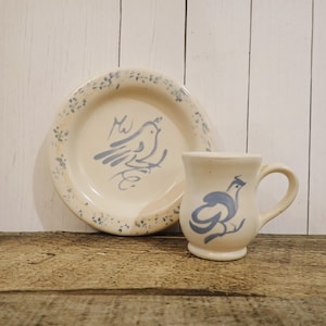 May include: A ceramic plate and matching mug set in an off-white finish. Both pieces feature a hand-painted blue bird illustration. The plate has a speckled blue rim, while the mug has a curved handle and a rounded base with a blue bird design.