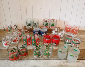 Puede incluir: Una colección de veintiocho vasos de vidrio transparente con diversos diseños navideños que incluyen árboles de Navidad, cascanueces, hojas de acebo, gansos y casas con el texto noel, dispuestos en varias filas sobre una superficie de madera.
