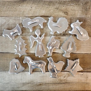 May include: A collection of twelve silver-colored metal cookie cutters in various shapes, including an angel, a bell, a star, a tree, a holly leaf, and several animal figures, each featuring a small metal handle attached to the top surface.