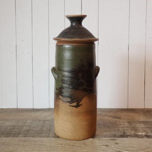 May include: A tall, handcrafted ceramic jar with a lid. The jar is cylindrical with a dark green and brown glaze, and has two small handles. The lid is a darker shade of brown. The jar is approximately 12 inches tall.