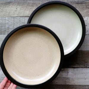 May include: Two round ceramic plates with a speckled beige interior and a thick black rim. One plate is a light beige color, and the other is a pale cream color. The plates are on a wooden surface.