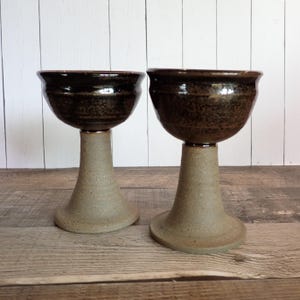 May include: Two handmade ceramic chalices. The bowls are dark brown with a glossy finish, and the stems are a light tan color. The chalices are displayed on a wooden surface with a white-painted wood background.