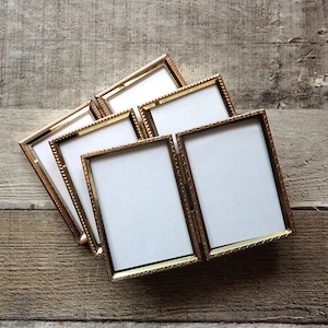 May include: A set of three hinged, rectangular picture frames made of gold-toned metal. Each frame features a textured, beaded edge detail and contains a blank white insert. The frames are connected by metal hinges, allowing them to fold.