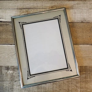 May include: Silver picture frame with a rectangular opening and a decorative black border. The frame has a silver finish and is set against a wooden background. The corners of the frame feature a stylized black design.