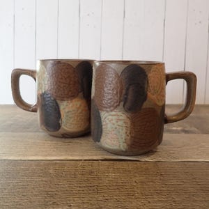 May include: Two ceramic mugs with a brown, tan, and black pattern. The mugs have a rectangular shape with a curved handle. The mugs are sitting on a wooden surface. The mugs are likely for coffee or tea.
