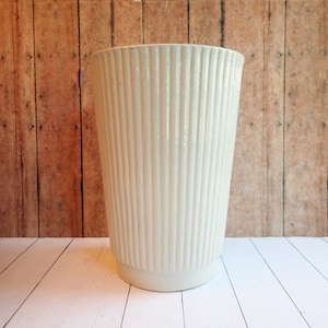 May include: A white ceramic planter with vertical ridges. The planter is sitting on a white wooden surface.