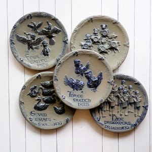 May include: Five circular ceramic plates featuring raised relief designs from the Twelve Days of Christmas. The plates depict Four Calling Birds, Three French Hens, Seven Swans Swimming, Nine Drummers Drumming, and a group of mice, all in blue and tan.