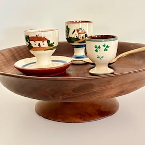 May include: A wooden pedestal serving tray holds three decorative egg cups. The egg cups are white with colorful painted designs, including houses and shamrocks. A gold-colored spoon rests on the tray.