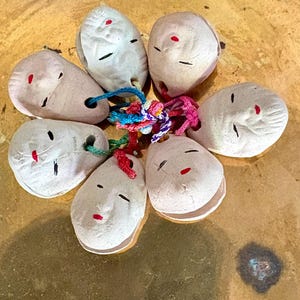May include: A cluster of six small, off-white ceramic mask charms arranged in a circular pattern. Each mask features simple, hand-painted black eyes and a red mouth. The charms are connected at the center by a bundle of multicolored braided cords.