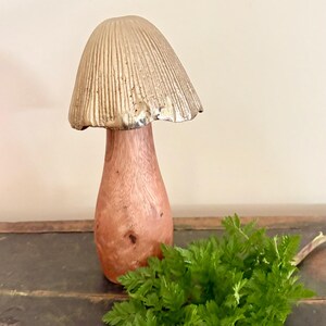 Pu&ograve; includere: Una scultura decorativa a forma di fungo con un cappello color oro e uno stelo in legno. Il fungo &egrave; posto su una superficie scura con un rametto di fogliame verde.