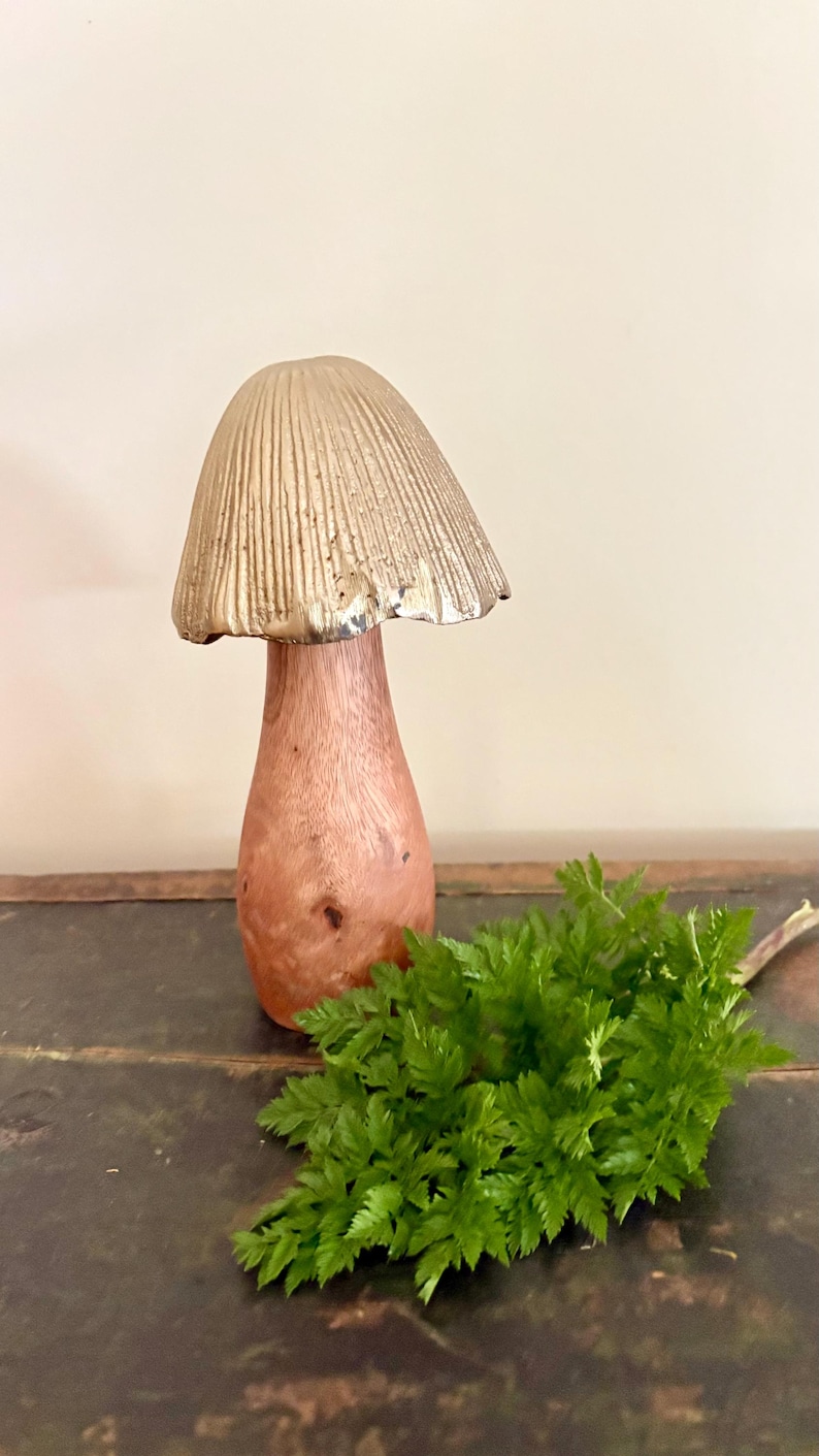 Pu&ograve; includere: Una scultura decorativa a forma di fungo con un cappello color oro e uno stelo in legno. Il fungo &egrave; posto su una superficie scura con un rametto di fogliame verde.