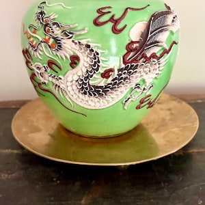 May include: A light green ceramic vase featuring a raised, detailed dragon motif in white, black, and red. The dragon wraps around the body of the vase, which has a smooth, glossy finish. The vase is resting on a circular, metallic gold-toned stand.