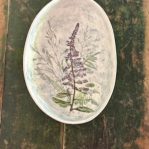 May include: An oval-shaped ceramic dish with a light blue and white mottled background. The center features a detailed botanical print of a purple flowering stalk surrounded by green fern fronds and thin, wispy grass-like stems.