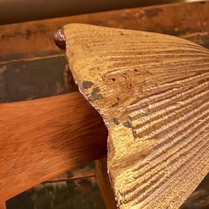 Pu&ograve; includere: Una sezione di un oggetto dorato a forma di ventaglio con una superficie strutturata. L'oggetto ha un manico in legno. La forma a ventaglio presenta una serie di linee parallele che si irradiano dal centro.