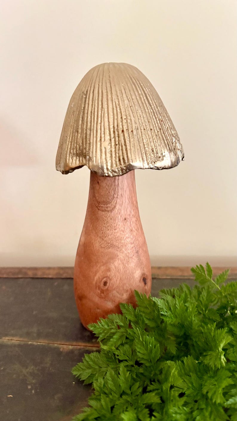 Pu&ograve; includere: Una scultura decorativa a forma di fungo con un cappello dorato e strutturato e uno stelo in legno. Il fungo &egrave; posto su uno sfondo neutro, con fogliame verde in primo piano. Un oggetto di arredamento per la casa unico.