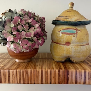 May include: A round wooden bowl filled with pink and green hydrangea flowers next to a yellow ceramic jar with a lid. The jar features a dark blue rim, a teal eye-shaped accent, and a rounded base. Both items sit on a shelf with a striped wood pattern.