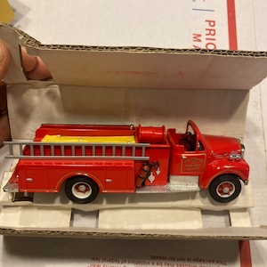 Puede incluir: Un modelo de camión de bomberos rojo con una escalera amarilla, neumáticos blancos y detalles plateados. El camión tiene las palabras "Fire Dept" en el lateral. El modelo está embalado en una caja de cartón.