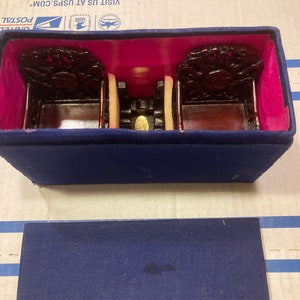 May include: A blue velvet box with a pink interior contains two small, intricately carved wooden chairs. The chairs are dark brown with gold accents and feature a dragon design. The box is closed and sitting on a blue surface.